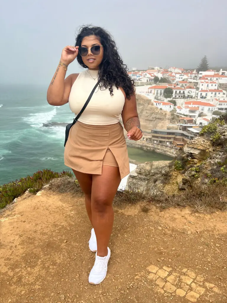Paige Savage standing on the Portugal coastline in a beige mini skirt and white tank top, overlooking the ocean and cliffside village.