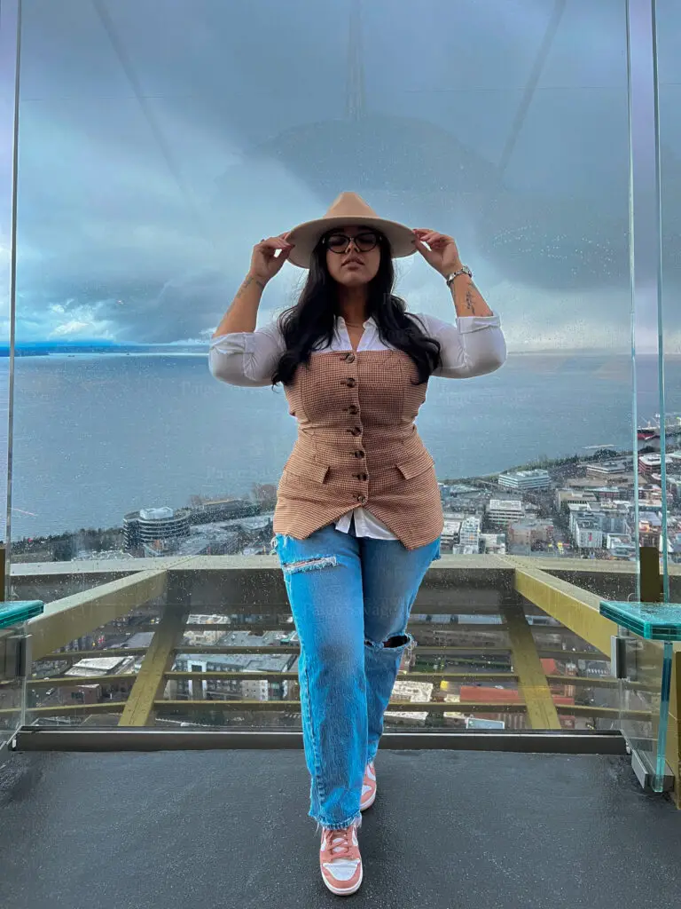 Paige Savage looking stylish, stands confidently on a glass observation deck on the Space Needle in Seattle overlooking the city skyline and water, dressed in a beige wide brim hat, glasses, brown corset vest, white blouse, ripped Levis jeans, and pink nike dunks. This "fly me to you" moment captures effortless cool with a moody sky backdrop.