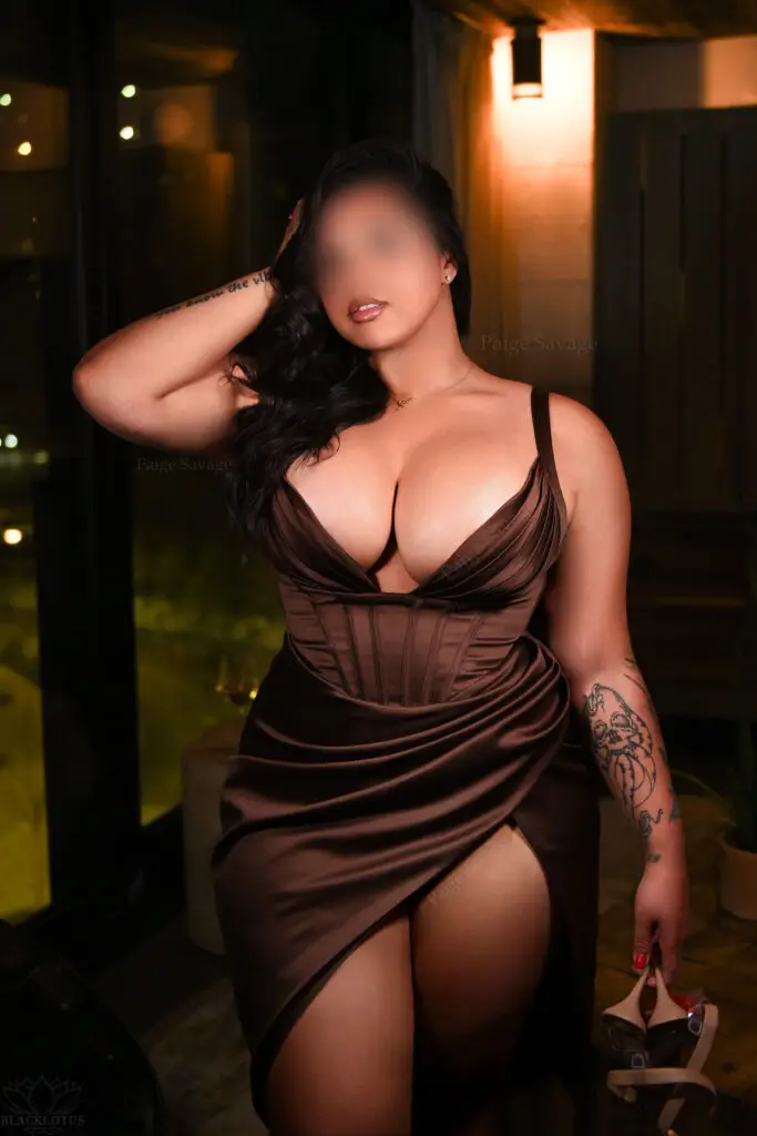 Paige Savage in a brown satin gown with a thigh-high slit, holding her heels and posing near a window overlooking DUMBO at night.