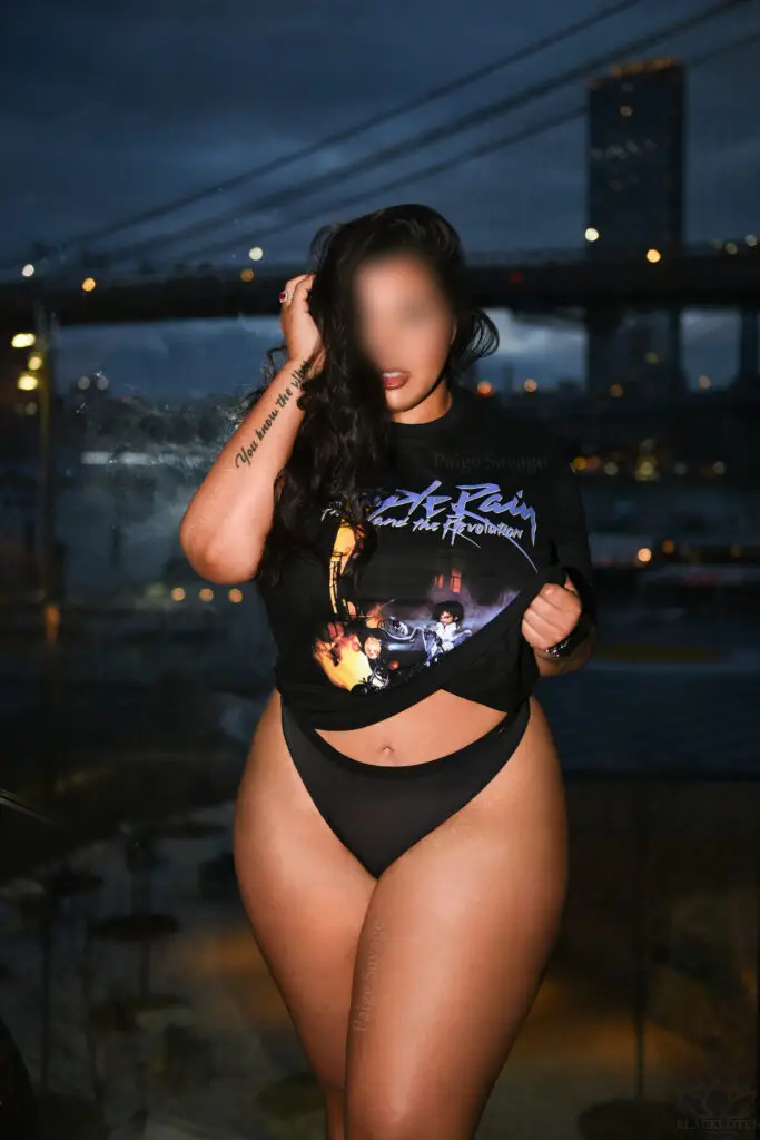 Paige Savage wearing a Prince t-shirt and black panties, posing by a window with the NYC skyline and Brooklyn Bridge behind her.