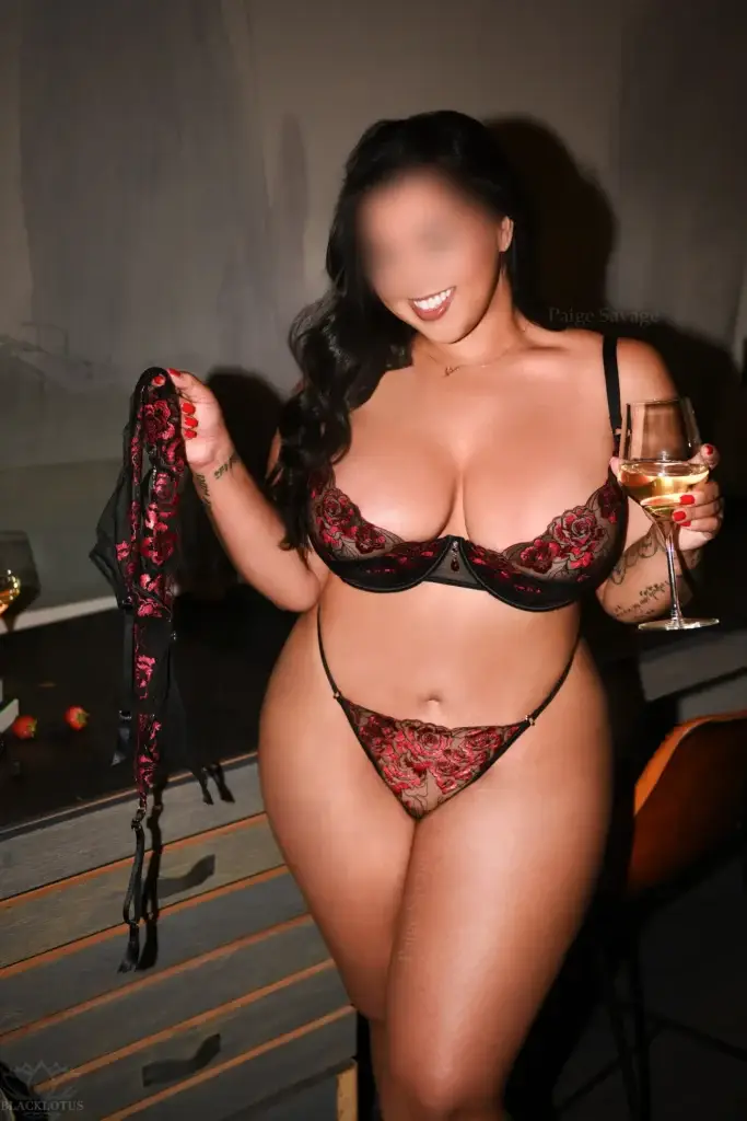 Paige Savage smiling in read and black lace lingerie, holding a glass of white wine and matching suspender in a NYC apartment setting.